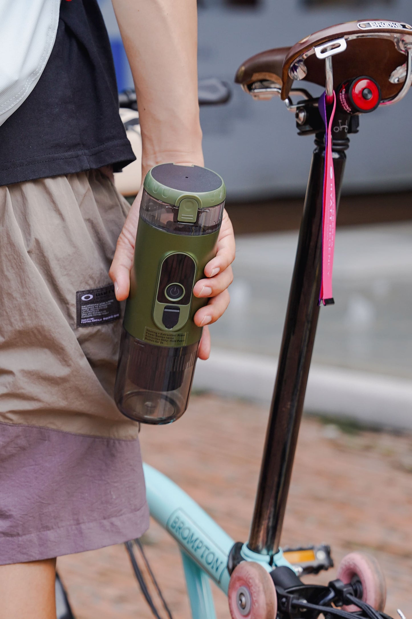 Hand holding TrailBrew green portable espresso maker beside a folding bicycle on urban street